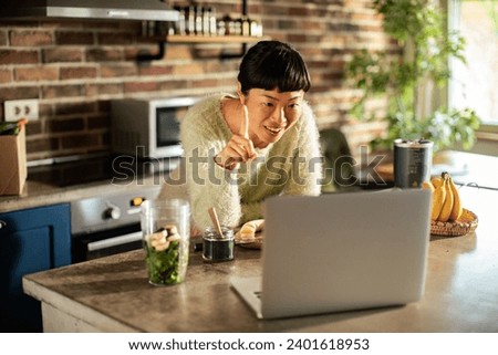 Asian Woman Preparing a Green Smoothie while Video Calling on Laptop in a Modern Kitchen Royalty-Free Stock Photo #2401618953