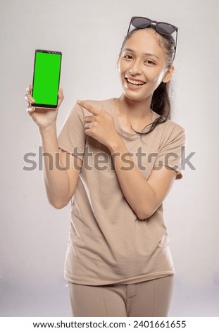 Beautiful Asian woman showing her cell phone screen while pointing at green screen and laughing happily in front of a white background.