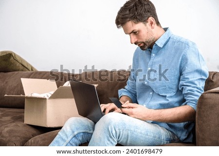 Adult man sitting on couch near opened box, looking on notebook and holding credit card in his hand. Contact with delivery company to complain on getting wrong order. Online shopping Royalty-Free Stock Photo #2401698479