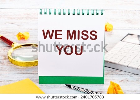 WE MISS YOU text on the white sheet of the notebook is next to a magnifying glass, calculator, pencil, stickers