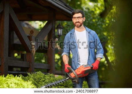 Attractive gardener posing, while trimming conifer bush by electric hedge clippers in sunny day. Portrait of male worker smiling at camera, while trimming thuja in garden. Concept of seasonal work.  Royalty-Free Stock Photo #2401708897