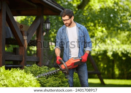 Dark haired gardener trimming overgrown green conifer bush by electric hedge clippers in summer. Front view of handsome man cutting thuja in garden, with blurred background. Concept of seasonal work.  Royalty-Free Stock Photo #2401708917