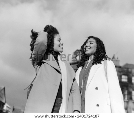 Two female black friends having fun in analog film monochrome portrait. They are outdoors, in the street, in a very cold winter day. They are smiling and looking each other.