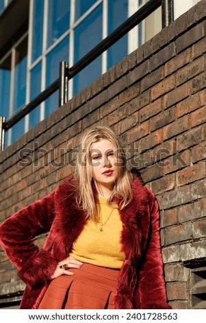 Hipster young woman posing against a brick wall with attitude. She looks really confident and she is wearing vintage outfits. Red faux fur coat, yellow sweater and orange skirt.