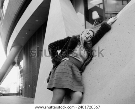 Monochromatic image of female model leaning on a modern building. Grainy image made with an analog medium format camera. Proud attitude and she is wearing vintage thick winter outfits.