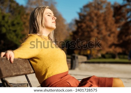 Sweet young woman sitting on a bench enjoying the sun in an autumn winter day. She is wearing and orange sweater and orange wool skirt. Relax and peace of mind concept.