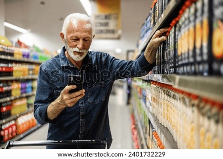 Senior Man doing grocery shopping at the supermarket, he is using apps on his smartphone. Male customer shopping with smartphone checklist, taking products from shelf at the shop.