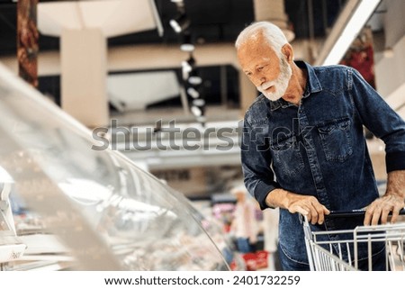 Portrait of mature Caucasian man doing grocery shopping at the supermarket. Smiling man pushing a shopping cart in the grocery store and choosing the products.