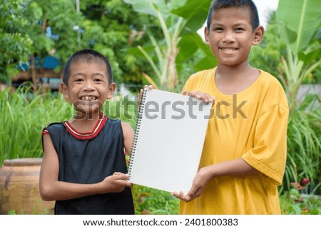 Two boys are smiling charmingly and holding a blank piece of paper or a sign on which to add words or letters in natural forest background at garden for concept of billboard.