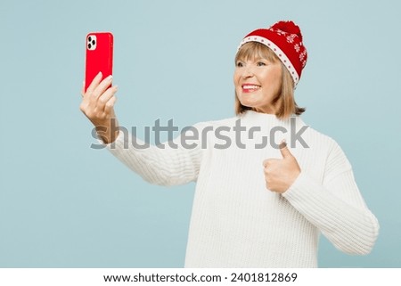 Merry elderly woman 50s years old wear sweater red hat posing doing selfie shot mobile cell phone show thumb up isolated on plain blue background. Happy New Year celebration Christmas holiday concept