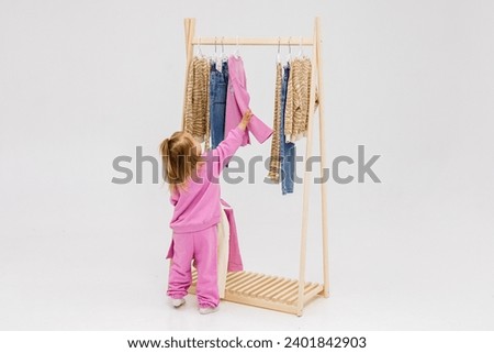 A child, a little girl, stands near the closet, chooses clothes against a light background. Dressing room with clothes on hangers. Wardrobe of children's and stylish clothes. Montessori wardrobe. Royalty-Free Stock Photo #2401842903