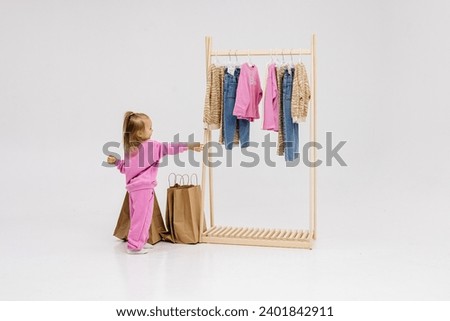 A child, a little girl, stands near the closet, chooses clothes against a light background. Dressing room with clothes on hangers. Wardrobe of children's and stylish clothes. Montessori wardrobe. Royalty-Free Stock Photo #2401842911