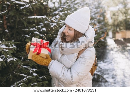 Surprised young 30s woman holding red gift box and get shock by open mouth on winter street. Winter holidays sale. Concept of shopping, holidays and happiness Christmas. High quality photo