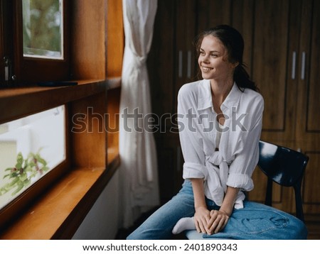 Woman sitting at home by a wooden window with a smile on a chair in homemade comfortable clothes and looking at the landscape, spring mood, women's day, rest on the weekend. Royalty-Free Stock Photo #2401890343