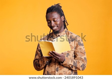 Cheerful african american man reading fiction book, enjoying learning new information standing in studio over yellow background. Clever person studying literature, liking genre and plot