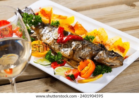 Picture of tasty baked whole trout with potatoes, greens and tomatoes on white plate, glass of wine