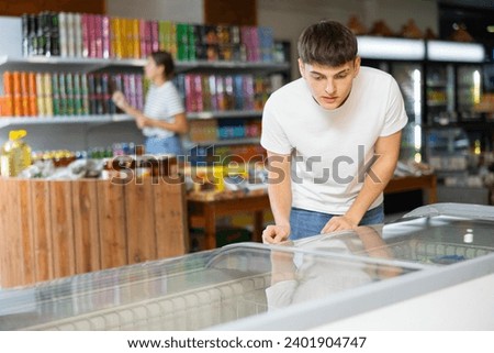 Young guy looks and chooses frozen food in refrigerator in supermarket. High quality photo