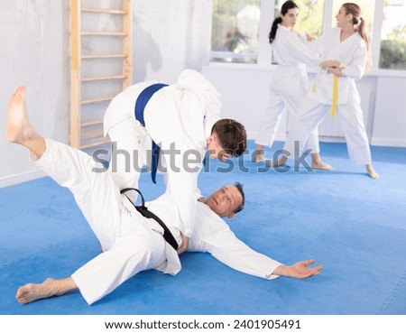 Adult man and young guy in kimonos train judo techniques in group in studio..