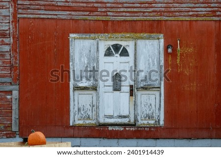 The exterior wall of an old red wooden shed. There's a white door with a semi circle window over the narrow entrance. A wooden decorative birdhouse hangs on the door. A pumpkin lays on the step. Royalty-Free Stock Photo #2401914439