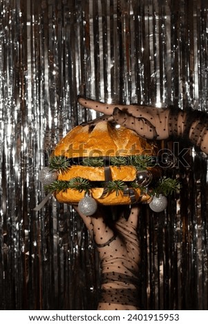 Vertical photo. Female hand in black tulle glove with polka dot pattern holds juicy tasty burger stuffed with fir branches and silver balls on silver striped background. Concept of Happy New Year 2024