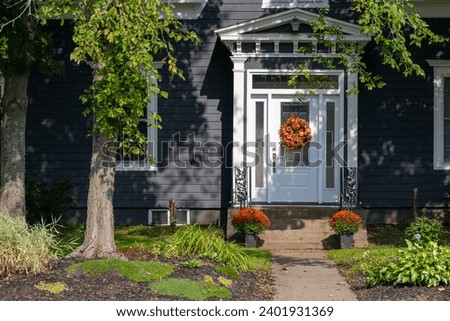 The entrance to a blue wooden historic house with a white door. There's an orange colored autumn decor wreath hanging on the door. Two mum flower pots are on the step. Trees and flowers cover the yard