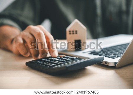 Pressing calculators hand eyes home refinance. Wooden house model, buy or rent note on desk. Saving for property purchase concept efficient mortgage payment. Tax credit analysis for financial strategy Royalty-Free Stock Photo #2401967485