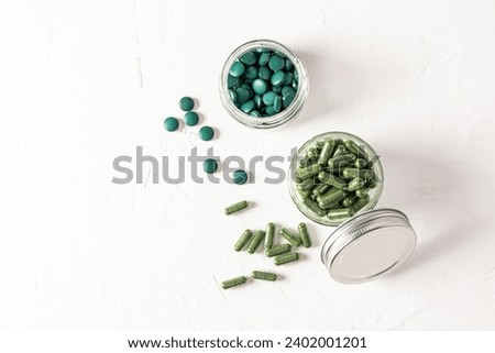Two glass jars filled with capsules and tablets of organic spirulina on a white textured background. Top view. Natural cleansing of the body Royalty-Free Stock Photo #2402001201