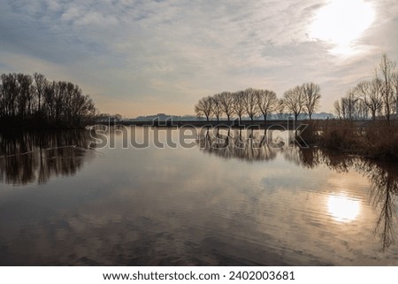 Landscape photo with a large lake and bare trees as silhouettes on the shore. It is a windless day and the water surface is very smooth. The low sun is just breaking through the cloudy sky.