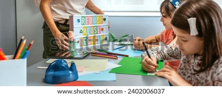 Unrecognizable female teacher holding lightbox with text reduce, reuse, recycle while young students drawing on colorful cardboards in ecology classroom. Sustainable lifestyle education concept.
