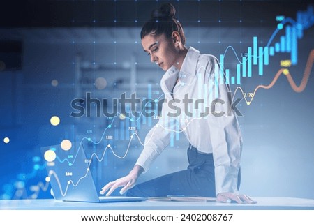 Attractive young businesswoman sitting on desk and using laptop with glowing candlestick forex chart on blurry office interior background. Trade, finance and stock concept