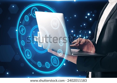 Close up of businessman hand using laptop with creative round medical interface with cross and other icons on blue background. Healthcare and innovation concept