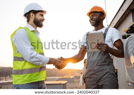Hardworking male workers doing hand shaking while signing online documents on personal tablet outdoors. Responsible air conditioning specialists making deal during work process on fresh air.
