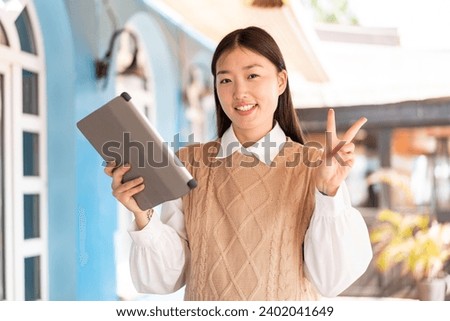 Young Chinese woman holding a tablet at outdoors smiling and showing victory sign