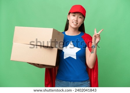 Super Hero delivery Asian woman holding boxes over isolated background smiling and showing victory sign