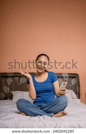 A beautiful Asian woman wearing a blue t-shirt sitting with her legs crossed on a bed using holding and using cell phone  while smiling cheerfully.