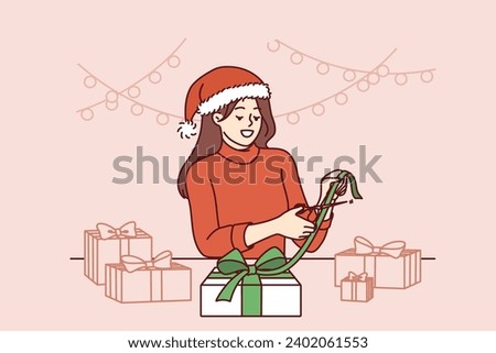 Christmas woman makes gifts with own hands, rejoicing at approach of new year holidays. Girl in santa hat preparing christmas giftbox for friends or family members and making bow from ribbon Royalty-Free Stock Photo #2402061553