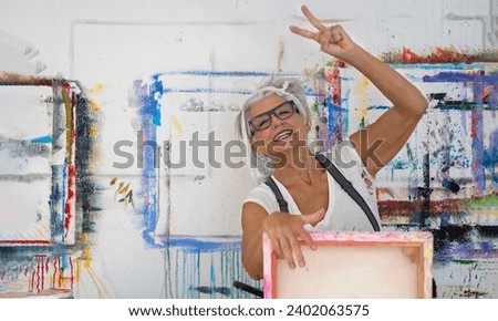 older gray haired mature happy laughing artist painter woman with glasses behind back of canvas painting stretches her arms upwards with power and joy, showing victory sign, in her studio, copy space