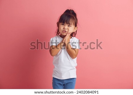 Funny little asian girl keeping palms pressed together in front of her, asks and begging something, has regretful or sorrowful expression standing over pink isolated background Royalty-Free Stock Photo #2402084091