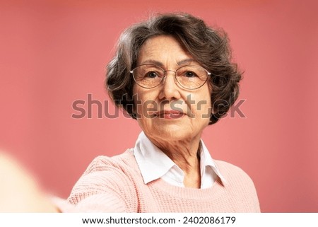 Portrait of serious attractive senior woman looking at camera, taking selfie, standing isolated on pink background. Advertisement concept Royalty-Free Stock Photo #2402086179