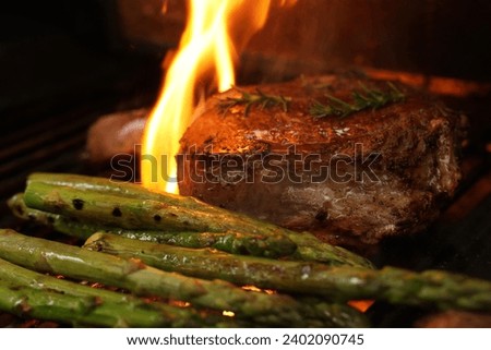 A mouth watering prefect bone-in rib-eye steak cooking on the bbq, barbecue, barbeque or griller with flames and asparagus sides. Rosemary garnish and close-up angle.  Royalty-Free Stock Photo #2402090745