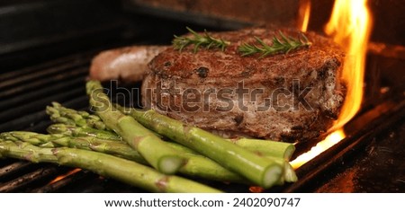 The prefect bone-in rib-eye steak cooking on the bbq, barbecue, barbeque or griller with flames and asparagus sides. Rosemary garnish and close-up angle.  Royalty-Free Stock Photo #2402090747