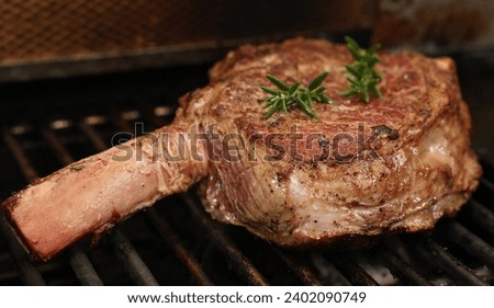 A single ribeye steak on the bone with rosemary herb garnish  in the early stages of cooking on the bbq or barbecue grill. The prefect cut of beef meat close up cooking on the griller. Royalty-Free Stock Photo #2402090749