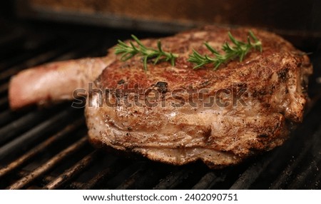 A single ribeye steak on the bone with rosemary herb garnish cooking on the bbq or barbecue grill. The prefect cut of beef meat close up cooking on the griller.  Royalty-Free Stock Photo #2402090751