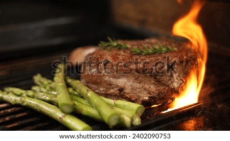 The prefect mouth watering bone-in rib-eye steak cooking on the bbq, barbecue, barbeque or griller with flames and asparagus sides. Rosemary garnish and close-up angle.  Royalty-Free Stock Photo #2402090753