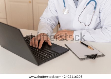 Male doctor working on his laptop in a medical office. Doctor in white uniform with stethoscope taking notes on PC writing in medical journal, professional therapist practitioner filling documents