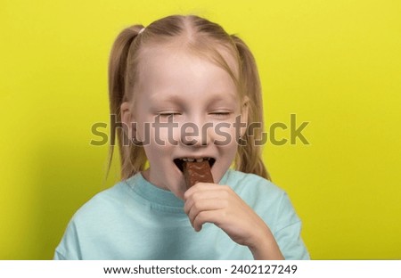 A beautiful joyful girl with white hair of seven years old eats a chocolate bar on a yellow background. Children are lovers of sweet junk food.