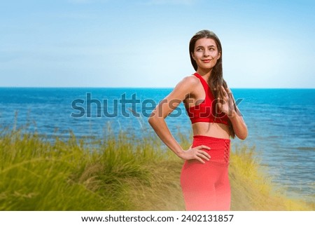 Portrait of happy fit young woman in red sportswear posing on beautiful beach, Seaside serenity: Young woman in sportswear finds relaxation by the ocean shore
