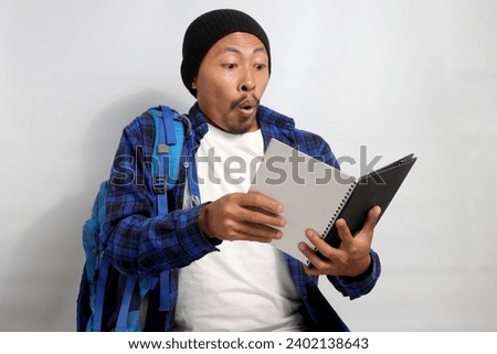 An excited young Asian student, dressed in a beanie hat and casual shirt, carrying a backpack, appears surprised while engrossed in reading a book against a white background