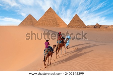 Camels in Giza Pyramid Complex - A woman in a red turban riding a camel across the thin sand dunes - Cairo, Egypt Royalty-Free Stock Photo #2402145399