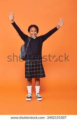 cheerful african american schoolgirl in uniform smiling and holding backpack on orange background Royalty-Free Stock Photo #2402155307